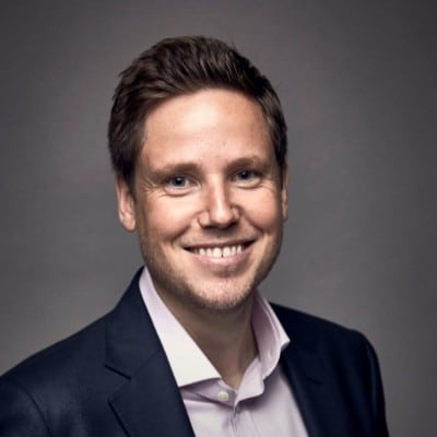 A man in a suit jacket and shirt smiles at the camera with a wide smile.