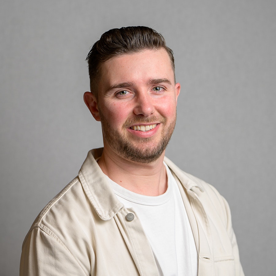 A man faces the camera wearing white and cream tops, he is smiling in front of a grey background