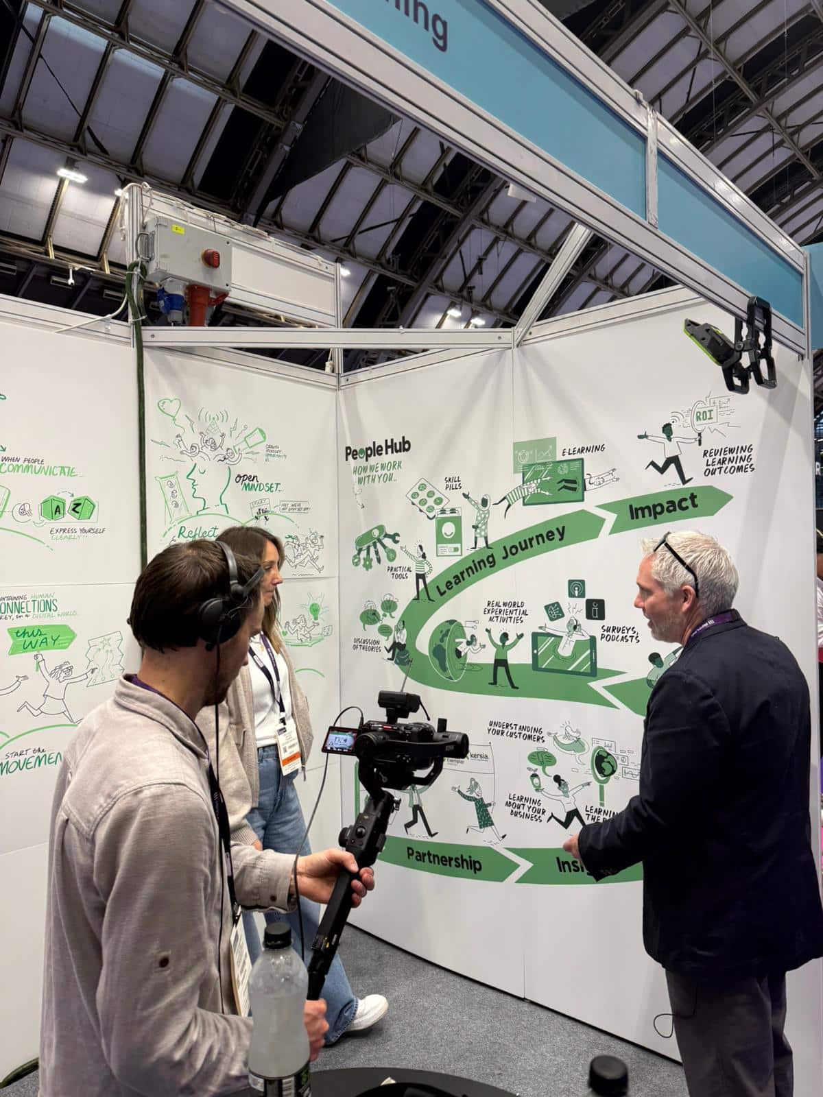A man holding camera equipment is filiming two men in front of a graphic at a trade show.