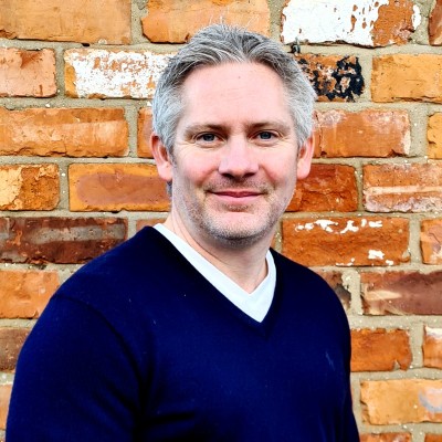 A man faces the camera. He is outside, smiling and in front of a brick wall.