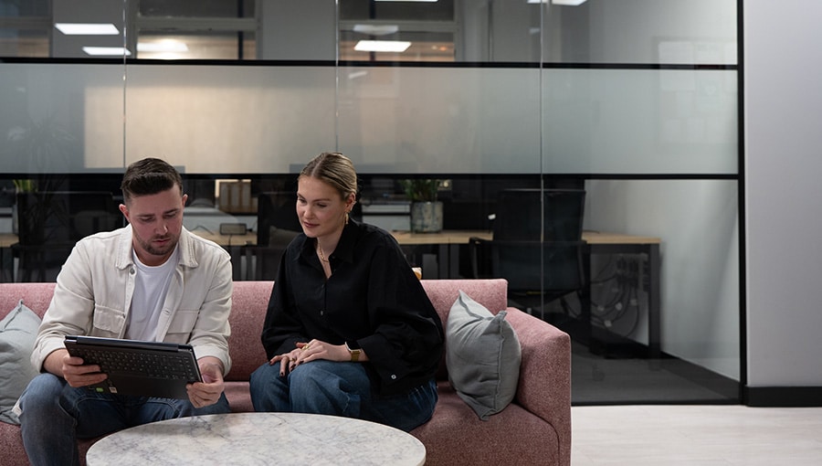 A man and a woman sit on a sofa in an office environment, it looks like they are working together.