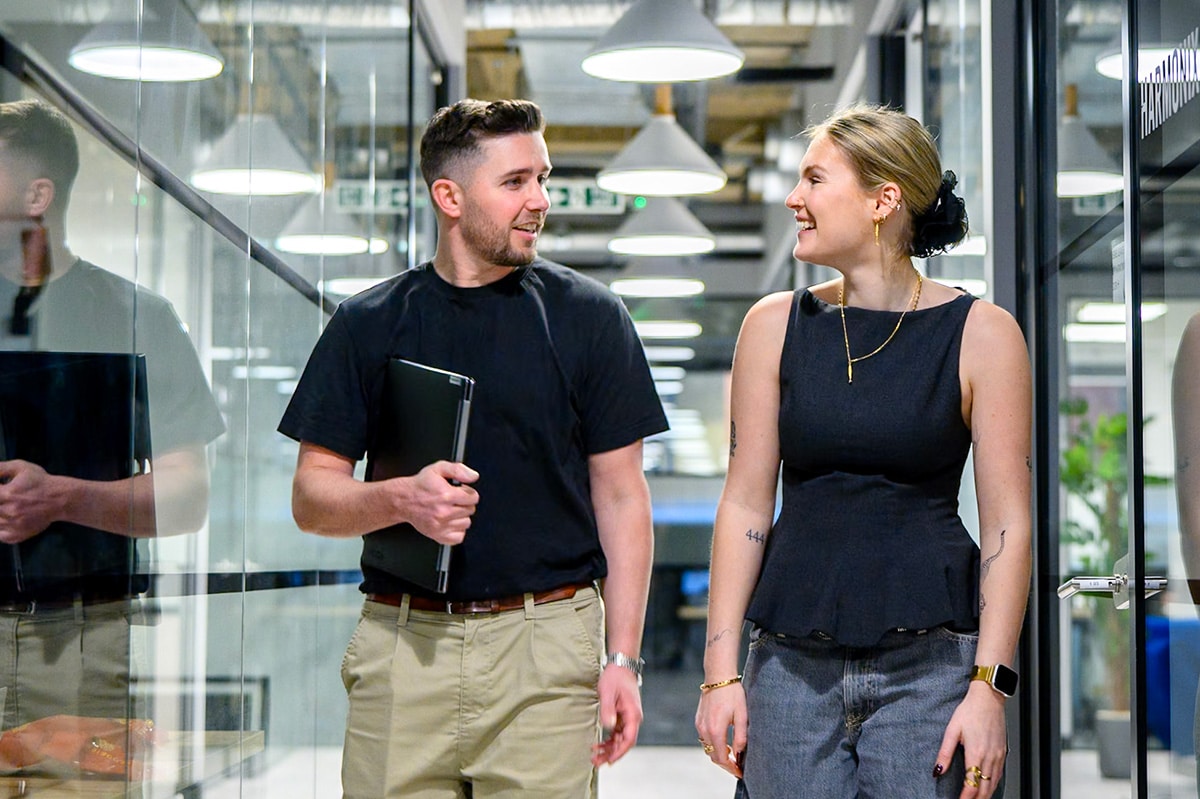 A man and a woman are smiling at each other while walking along a corridor in what looks like a modern office