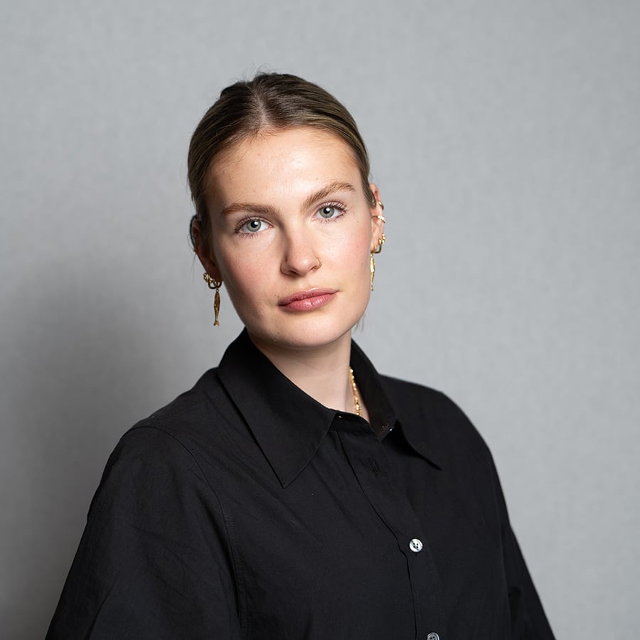 A woman with tied back hair in a black top faces the camera, with a grey background